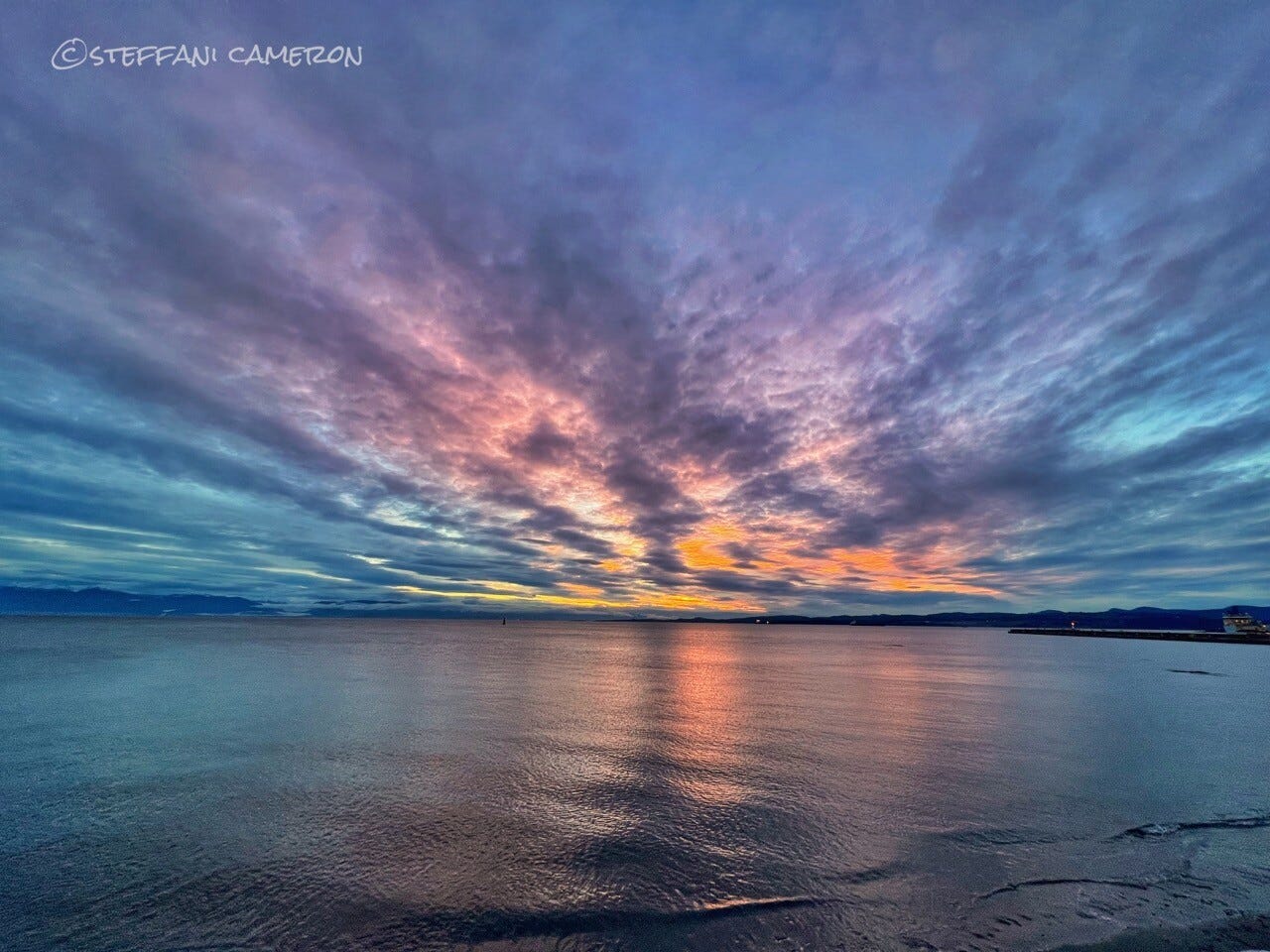 A partly cloudy sunset has wispy clouds radiating out from the sunset. There’s a pale blue sky darkening with the wispy clouds but the colours of the sunset are all smudged together — pink, yellow, orange, purple, white, and even green